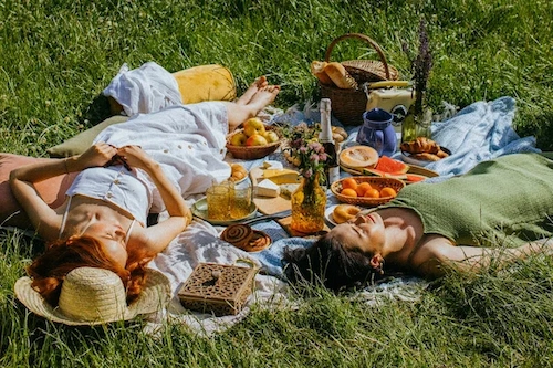 Zwei Frauen liegen entspannt auf einer Picknickdecke in einer hohen Sommerwiese neben einem Korb mit frischem Obst und Gebäck; finden Sie Ihre innere Balance mit Vitawy durch Achtsamkeit, Entspannung und Strategien für mentale Stärke.