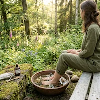 Eine Frau in grüner Leinenkleidung sitzt auf einer Holzbank im Wald und macht ein Fußbad in einer Tonschale mit Wasser und Kieselsteinen. Wir sind ein Team von passionierten Gesundheitsenthusiasten, die sich der Aufklärung und Inspiration verschrieben haben, um zu zeigen, wie die Kräfte der Umwelt die Selbstheilung aktivieren können.